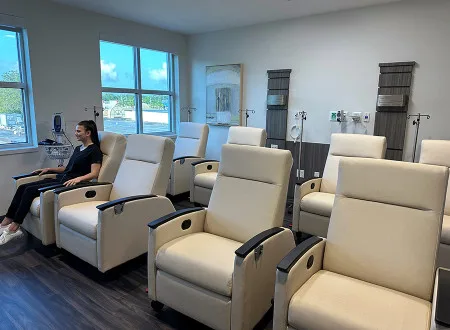 Wide view of a medical infusion center with rows of empty reclining chairs, IV equipment, and large windows providing natural light.