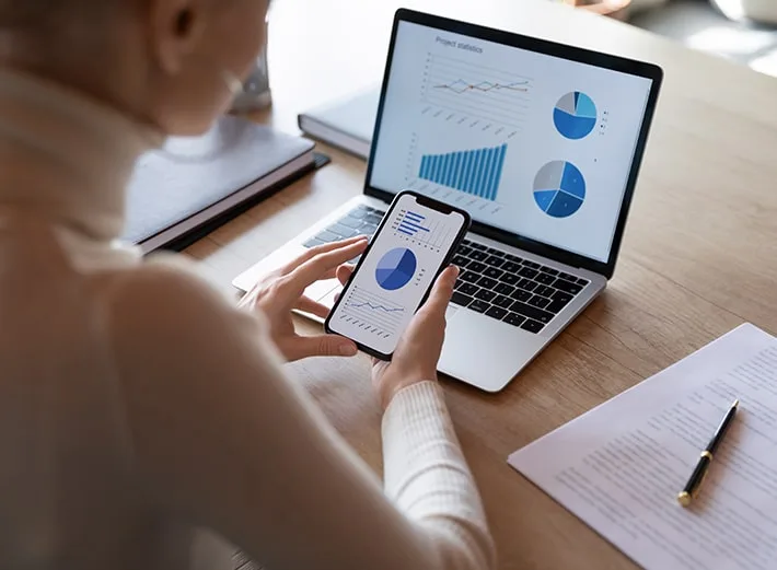 Person seated at a desk holding a smartphone while viewing charts and graphs on both the phone and a laptop, representing data analysis or performance metrics.