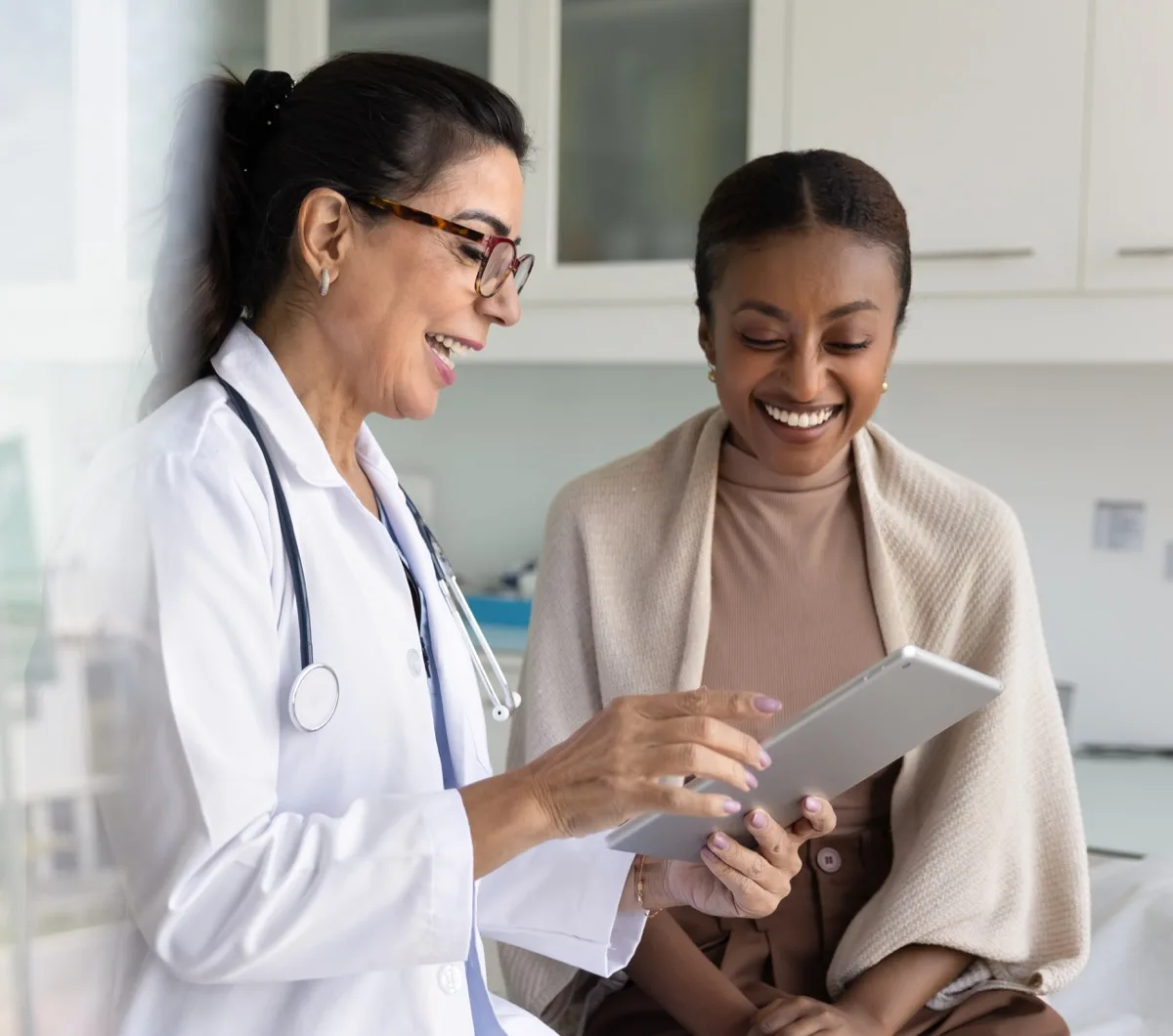 Happy senior practitioner woman and cheerful young African patient woman watching examination medical report on tablet computer, using digital technology, laughing