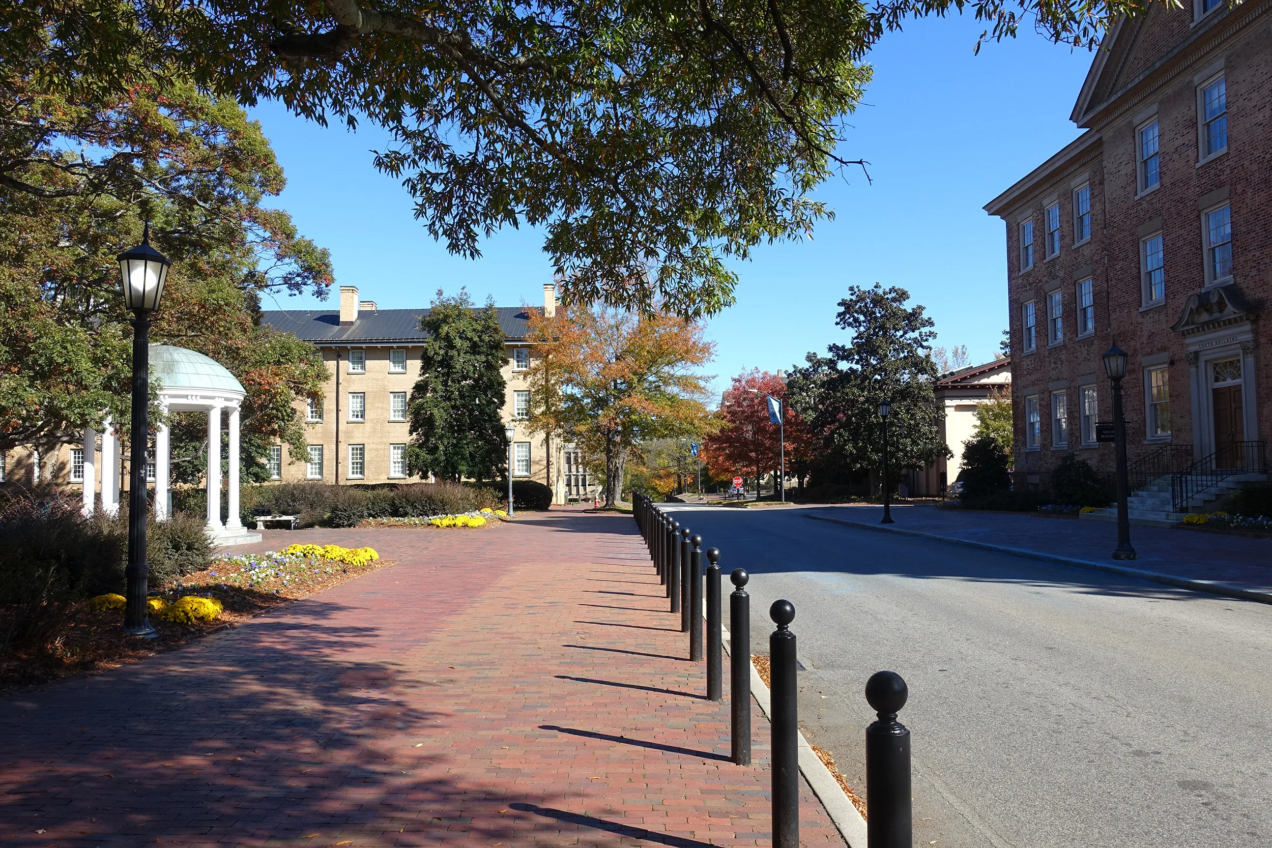 Brick walkway lined with black bollards leading through a university campus, with historic brick academic buildings on both sides, a white domed gazebo on the left, street lamps, and autumn trees with colorful foliage under a clear blue sky