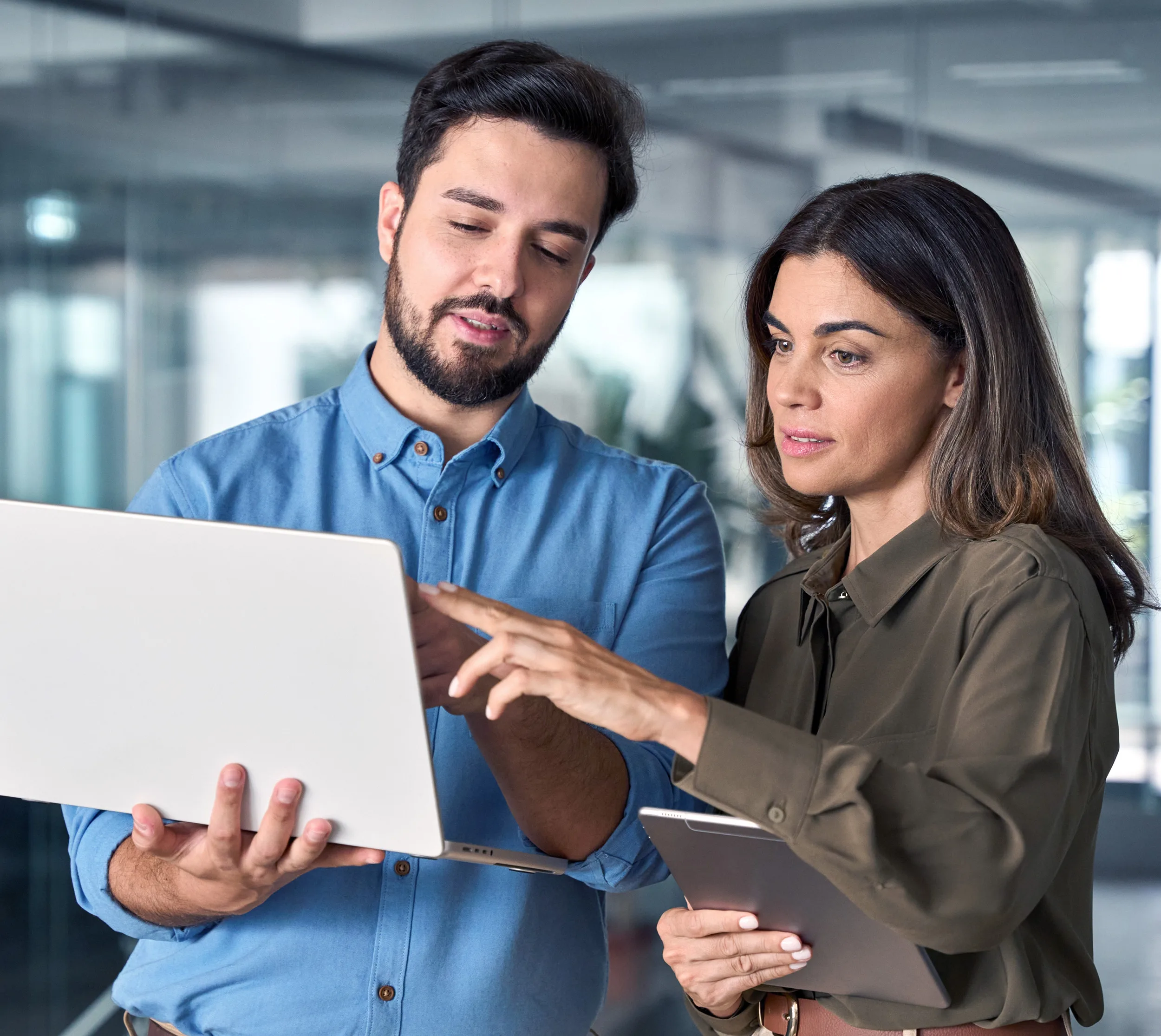 Two coworkers standing in an office environment, reviewing information on a laptop together while one person points at the screen and the other holds a tablet, suggesting collaboration or discussion of work tasks