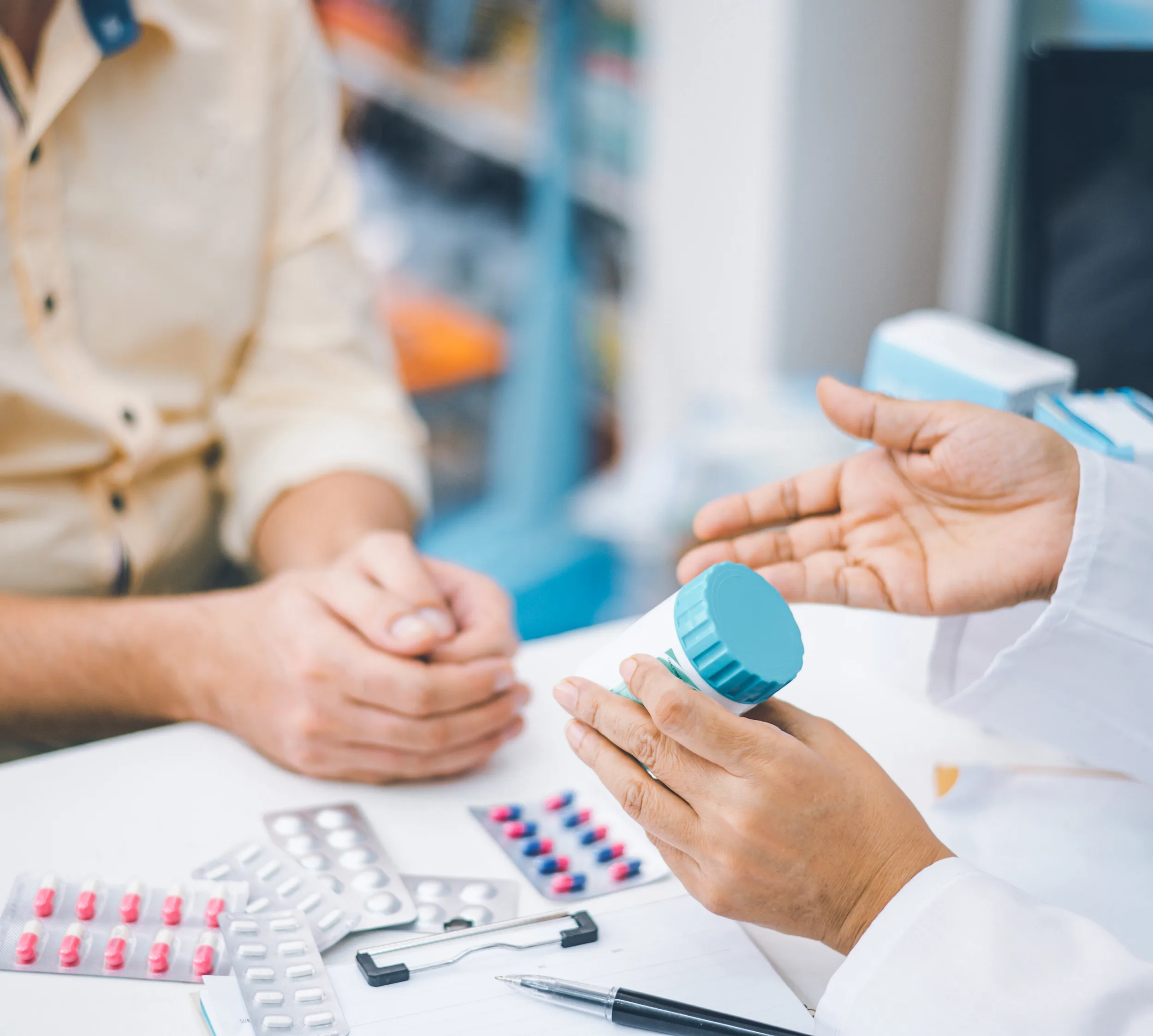 Pharmacist holding a prescription bottle and explaining medication to a patient across a counter, with blister packs of pills, a clipboard, and a pen visible on the table in a pharmacy setting